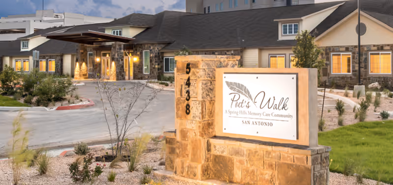 Exterior view of Poet's Walk San Antonio, a memory care community, showing the building entrance with warm lighting, a stone sign with the community name and address, and landscaped surroundings under a partly cloudy sky.