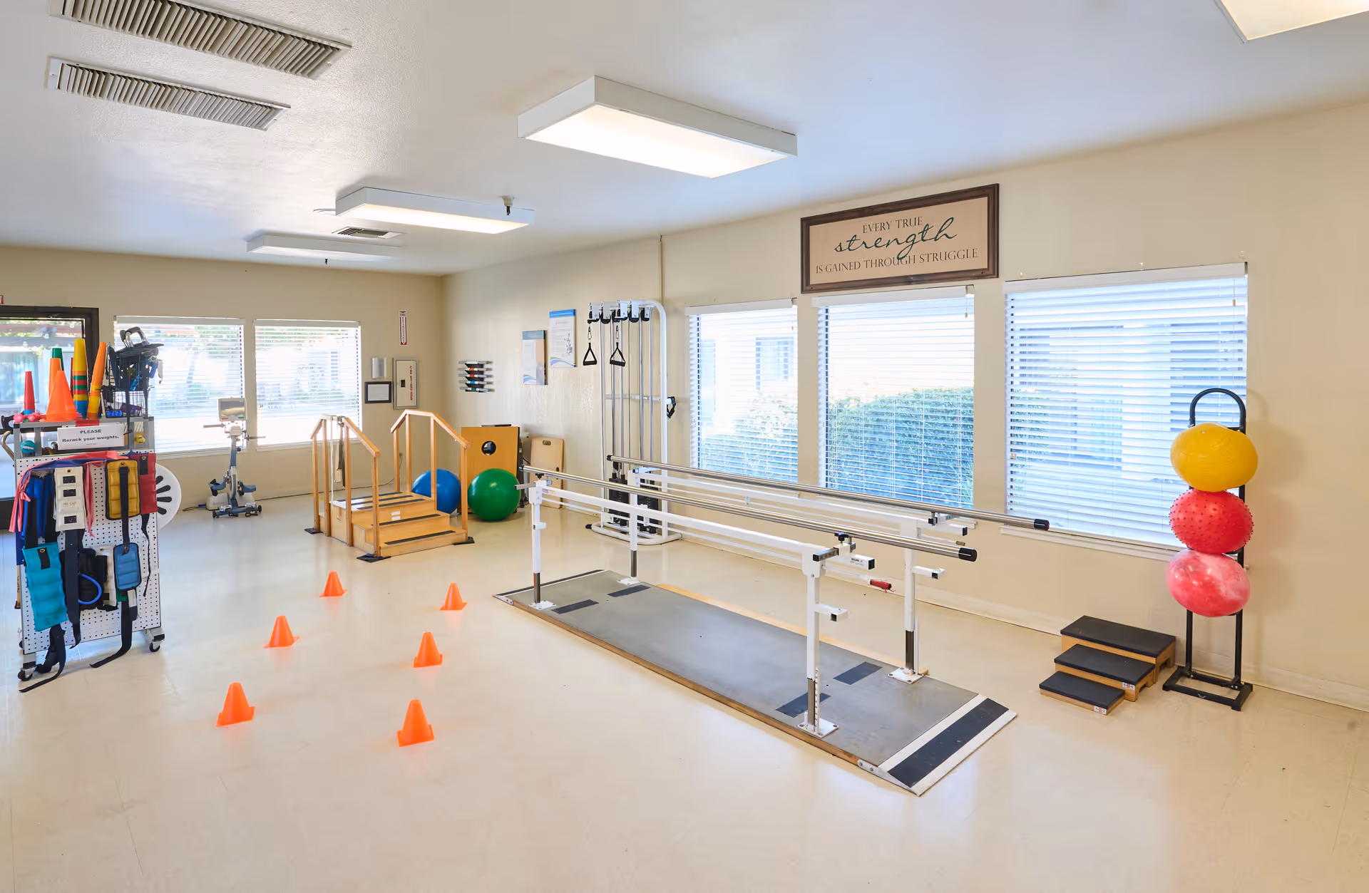 Bright rehabilitation exercise room with parallel bars, orange cones, therapy balls and assorted equipment by large windows.