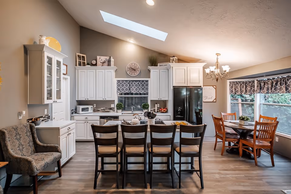 Open kitchen with a central island and four bar stools, white cabinets, skylight, and an adjacent dining table by large windows.