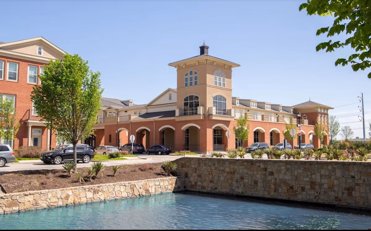 Exterior view of a senior living facility with a brick and beige building featuring arched windows and a tower-like structure. There are several cars parked in front, a stone wall bordering a water feature, and trees with green leaves under a clear blue sky.