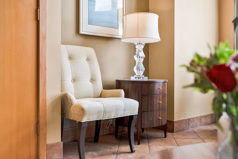 A cozy corner with a beige upholstered armchair next to a dark wooden side table with three drawers. On the table is a decorative lamp with a white lampshade. The floor is tiled and there is a framed picture hanging on the beige wall behind the chair. A blurred bouquet of flowers is partially visible in the foreground on the right.