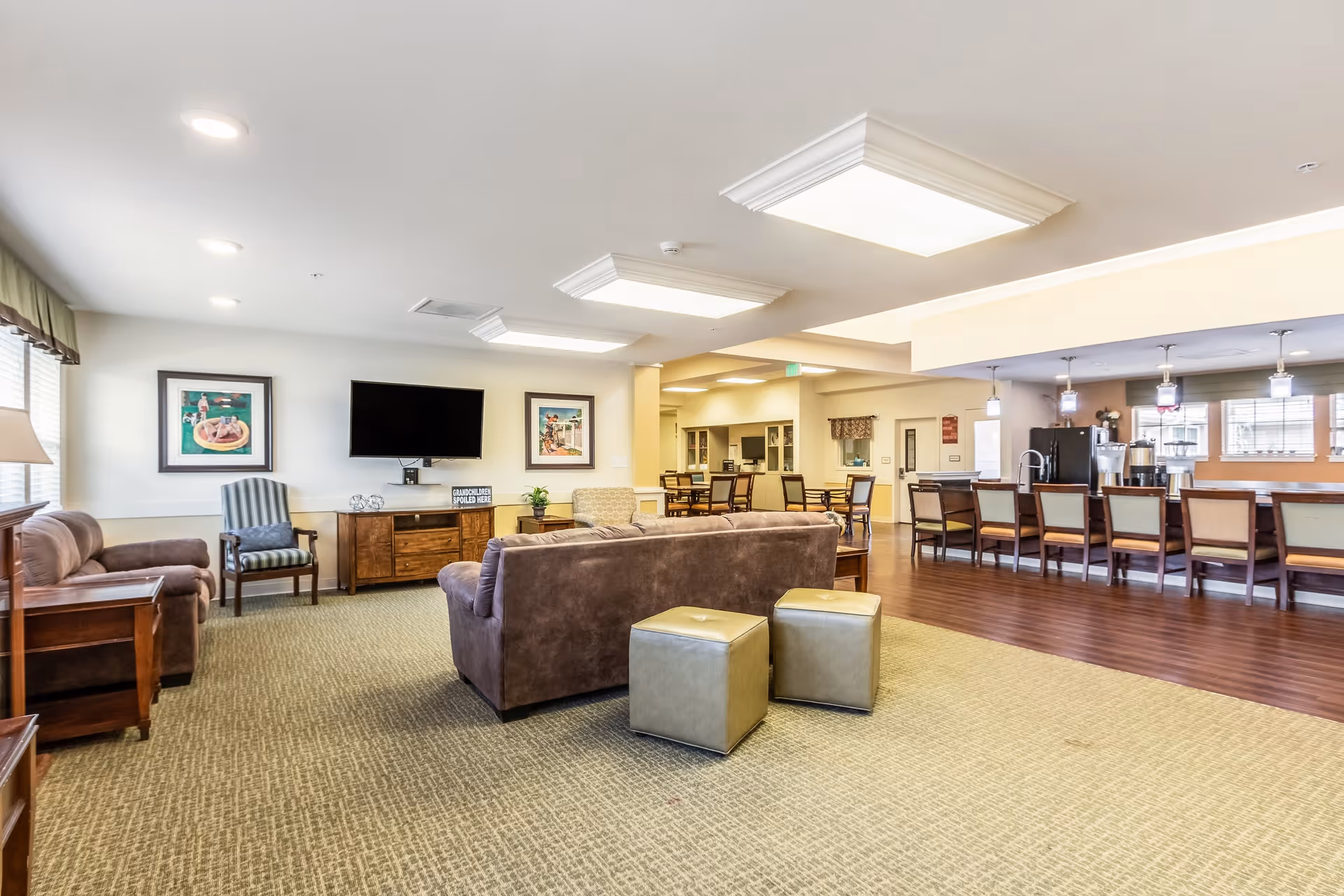 A spacious senior living common area featuring a seating arrangement with brown sofas, two small ottomans, and a striped armchair. The room has a wall-mounted flat-screen TV, framed artwork, and large windows with curtains allowing natural light. In the background, there is a dining area with tables and chairs, and a kitchen counter with bar stools and pendant lighting above.