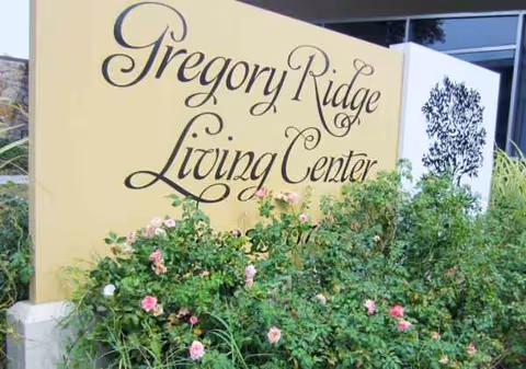 Beige building sign reads Gregory Ridge Living Center with flowering bushes in front.
