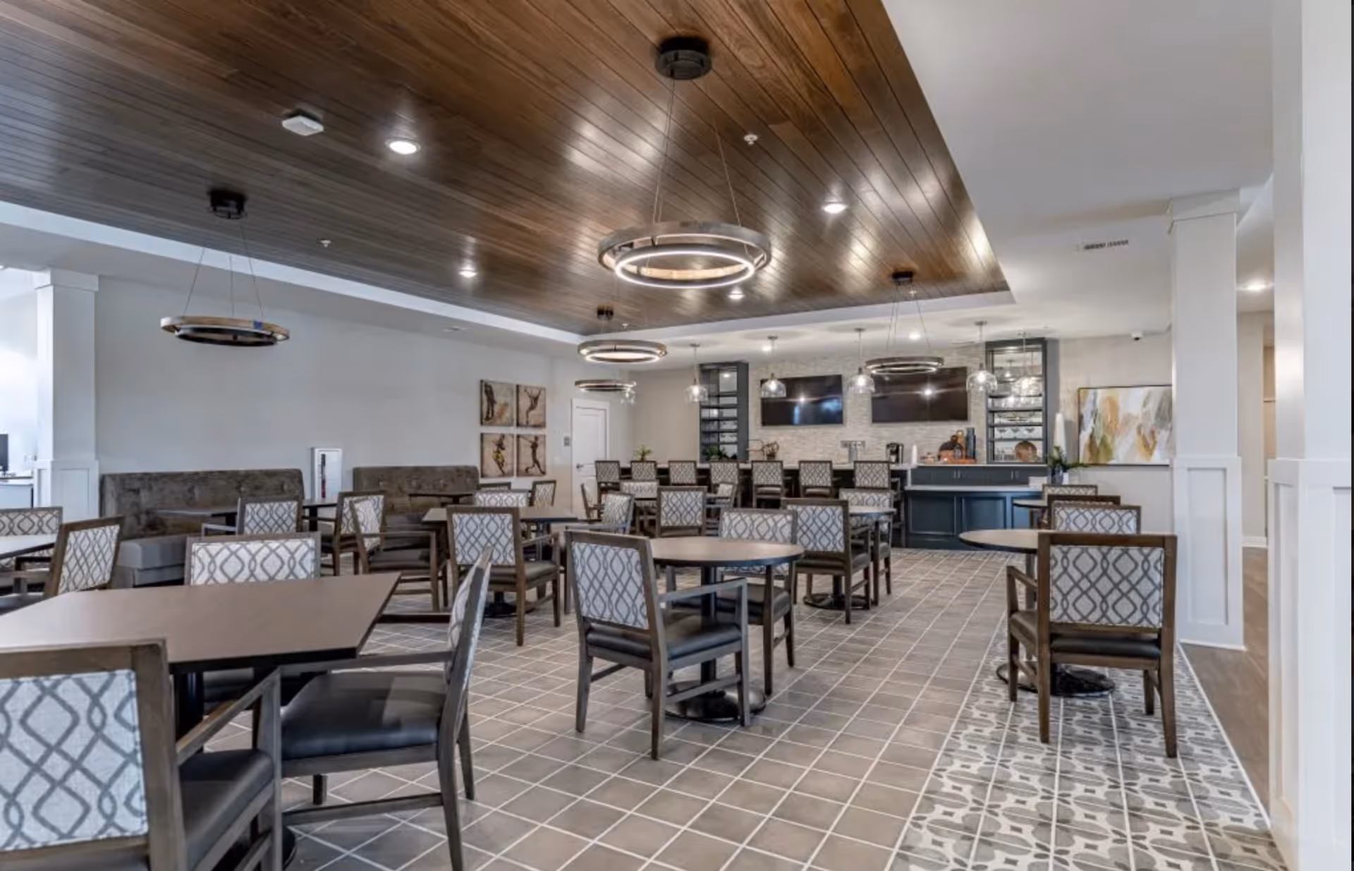 A spacious dining area in Avalon Senior Living featuring multiple tables and chairs with patterned upholstery. The room has a tiled floor with a decorative border, a wooden ceiling with modern circular light fixtures, and a serving counter with two large TVs mounted on the wall behind it. Artwork and plants decorate the space.