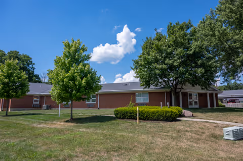 Single-story brick senior living building with a lawn, trees, and a walkway under a blue sky.