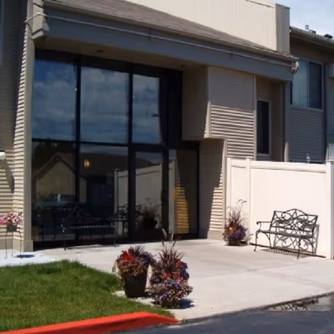 Outdoor patio area of Westgate Apartments with a concrete walkway, two metal benches, potted plants with flowers, and large glass windows reflecting the sky and nearby buildings.