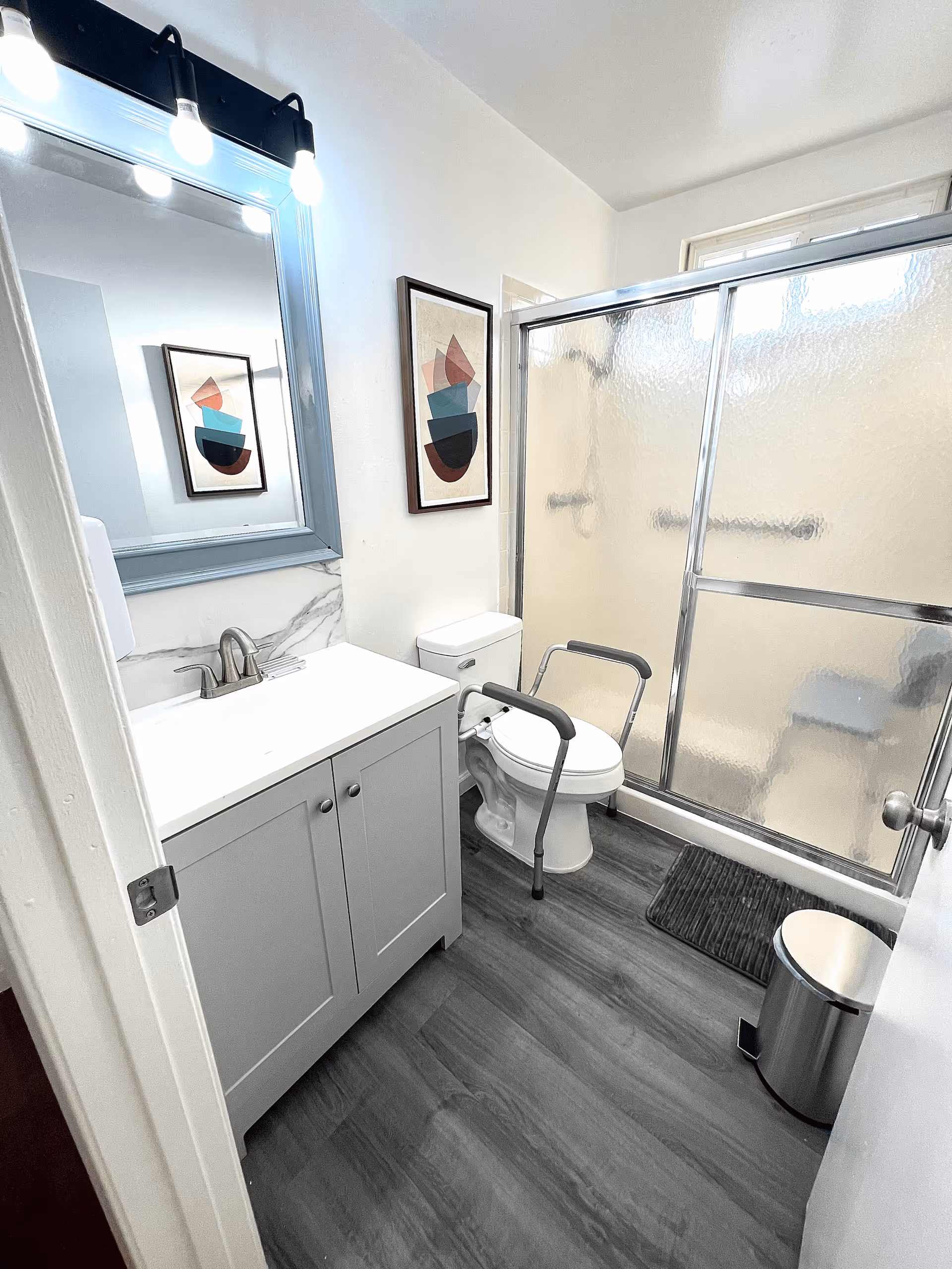 Small, bright bathroom with a vanity and mirror, a toilet fitted with safety grab rails, and a sliding glass shower door.