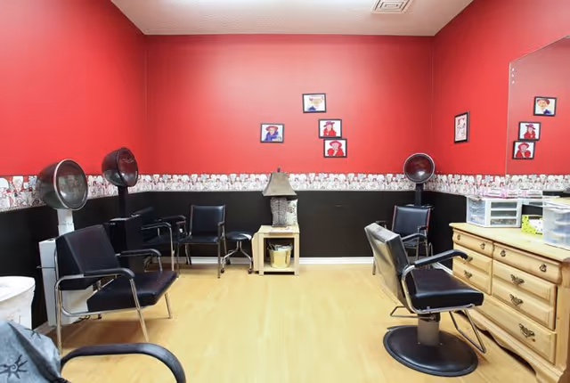 Interior of a salon room with red and black walls, featuring several black salon chairs, hair dryers, a wooden dresser with storage containers, a small table with a lamp, and framed pictures on the walls.