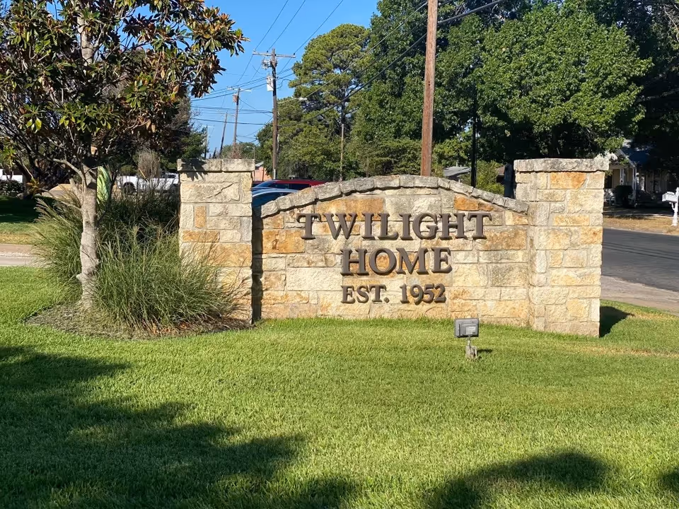 Stone sign in a grassy area with the text 'TWILIGHT HOME EST. 1952' surrounded by trees and a street in the background under a clear blue sky.