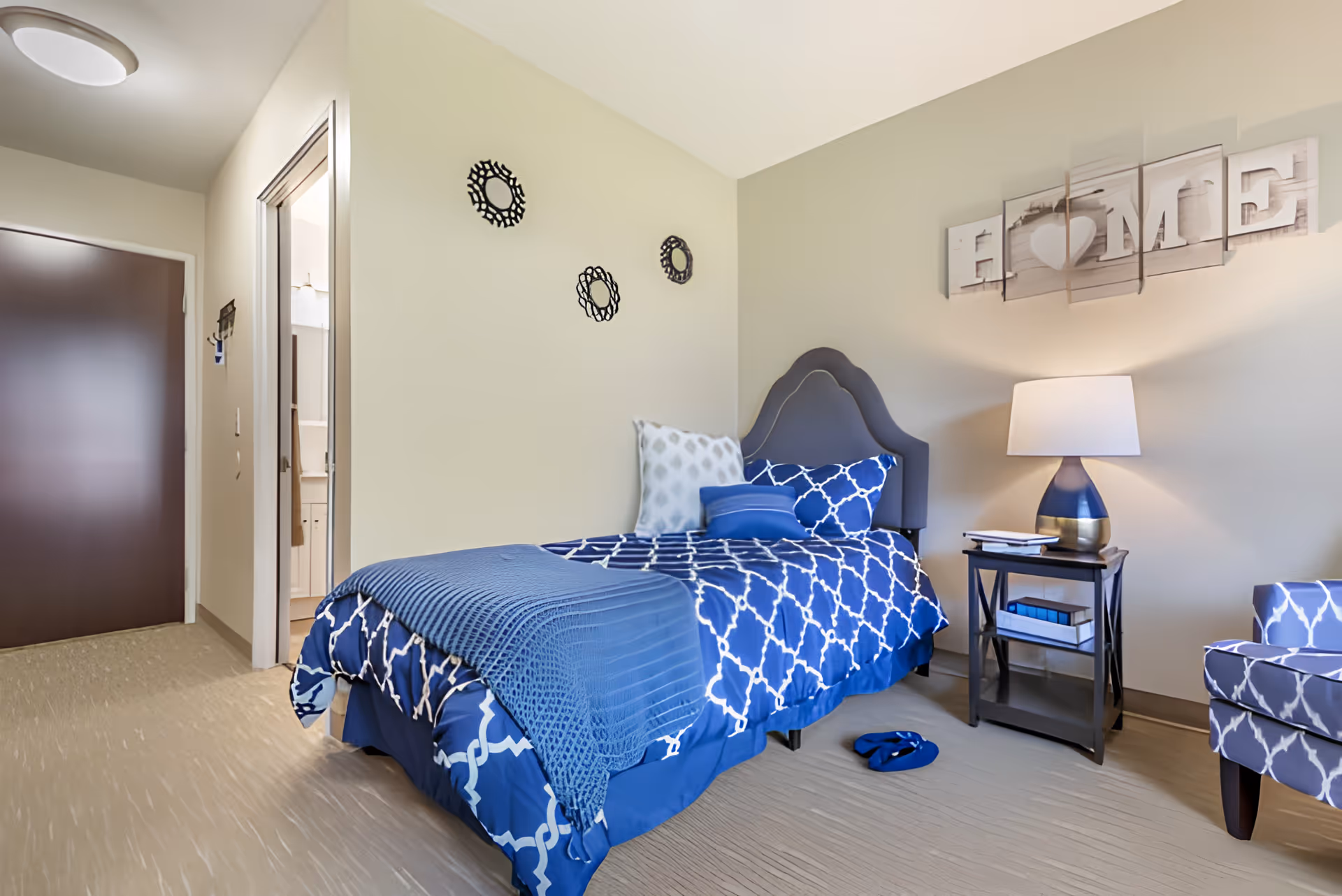 A neatly decorated bedroom with a single bed in blue patterned bedding, a nightstand with a lamp, and a matching armchair.