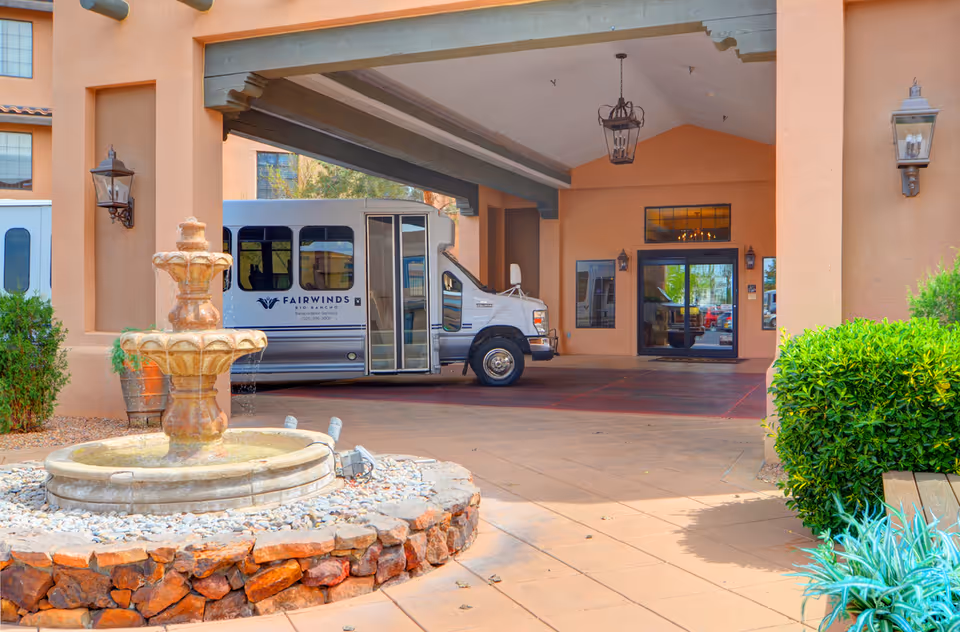Entrance area of Fairwinds - Rio Rancho facility with a covered drop-off zone. A silver shuttle bus with the Fairwinds logo is parked under the canopy. In the foreground, there is a stone fountain surrounded by rocks and greenery, with a tiled walkway leading to the glass entrance doors.