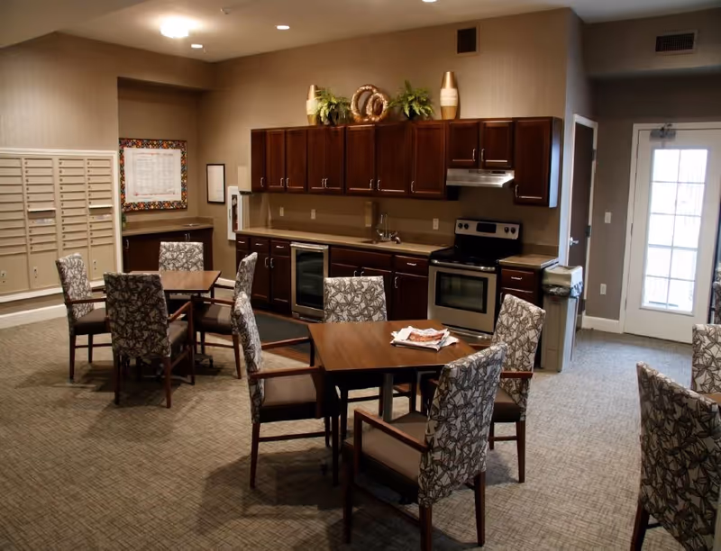 Interior view of a communal dining and kitchen area with several square wooden tables surrounded by patterned upholstered chairs. The kitchen features dark wood cabinets, a stainless steel stove, a small refrigerator, and decorative plants on top of the cabinets. There is a bulletin board and mailboxes on one wall, and a glass door leading outside.
