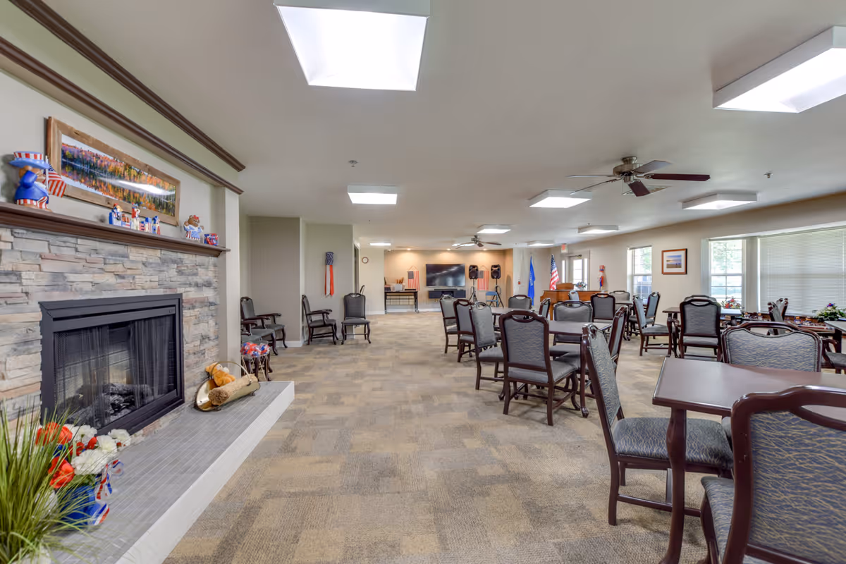 A spacious senior living community room with multiple tables and chairs arranged for social gatherings. The room features a stone fireplace decorated with patriotic items, a large flat-screen TV mounted on the far wall, ceiling fans, and large windows allowing natural light to fill the space.