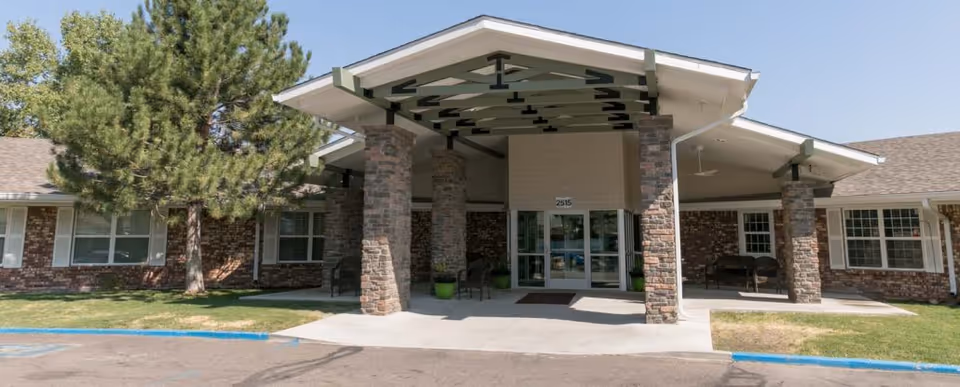 Front entrance of a brick rehabilitation center featuring a covered porte-cochere with stone columns, glass double doors, and adjacent lawn and trees.