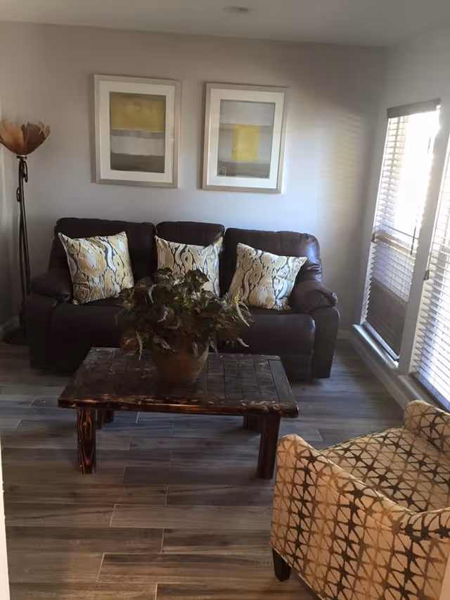 A cozy living room with a dark brown leather sofa adorned with three patterned cushions. In front of the sofa is a rustic wooden coffee table with a potted plant on top. To the right, there is a beige armchair with a geometric pattern. The room has light-colored walls with two framed abstract paintings hanging above the sofa. Large windows with blinds allow natural light to fill the space. A tall floor lamp stands in the corner to the left of the sofa.