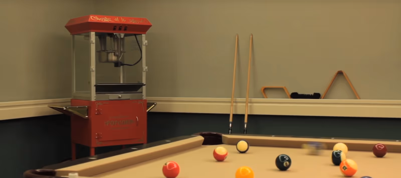 A recreational room corner featuring a red popcorn machine and a pool table with billiard balls scattered on its surface. Two pool cues are standing upright against the wall, and a rack for the cues is mounted on the wall behind the table.