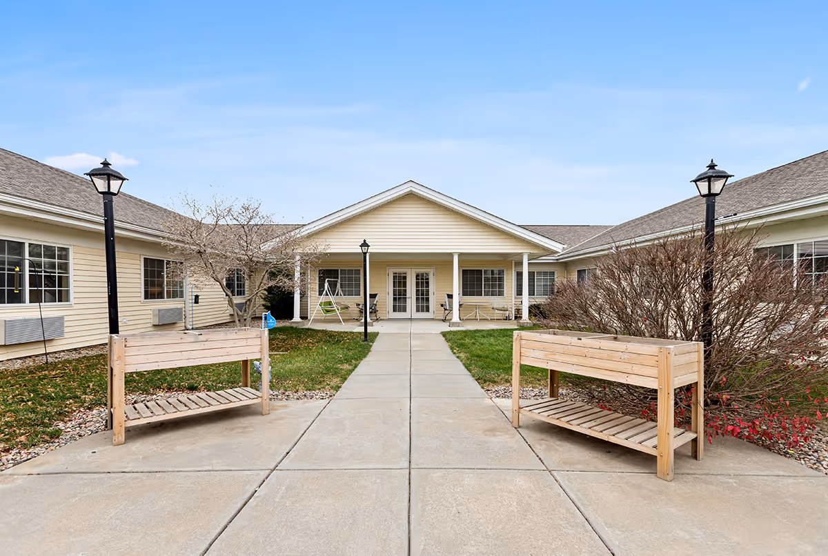 Outdoor courtyard area of a senior living facility with a concrete walkway leading to a covered entrance. The courtyard features two wooden benches, lamp posts, leafless trees, and bushes. The building has beige siding and multiple windows.