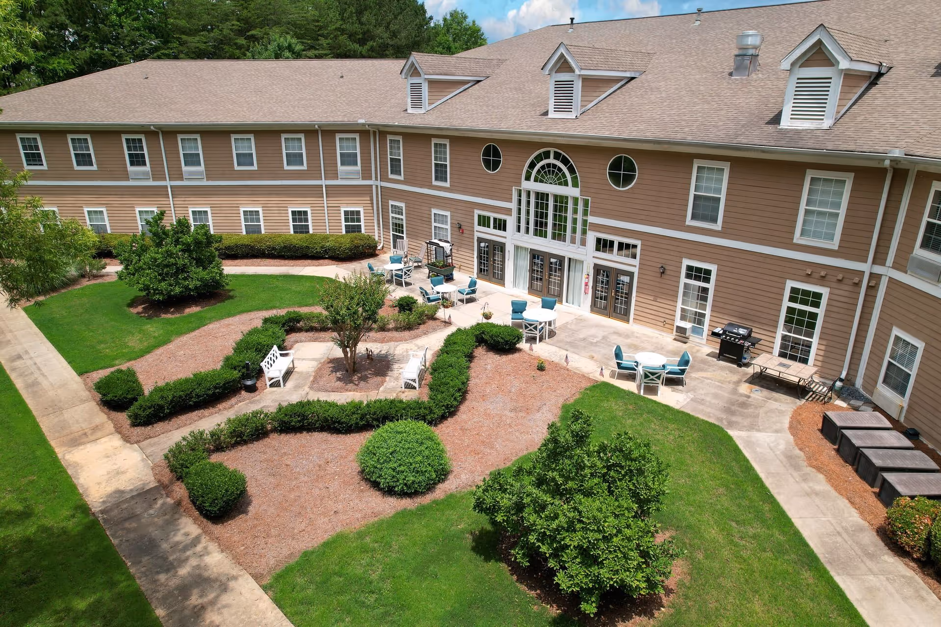 Outdoor courtyard area of a senior living facility with landscaped garden beds, bushes, and trees. There are several white benches and round tables with blue chairs on a concrete patio adjacent to a two-story building with multiple windows and a large arched window above double doors.