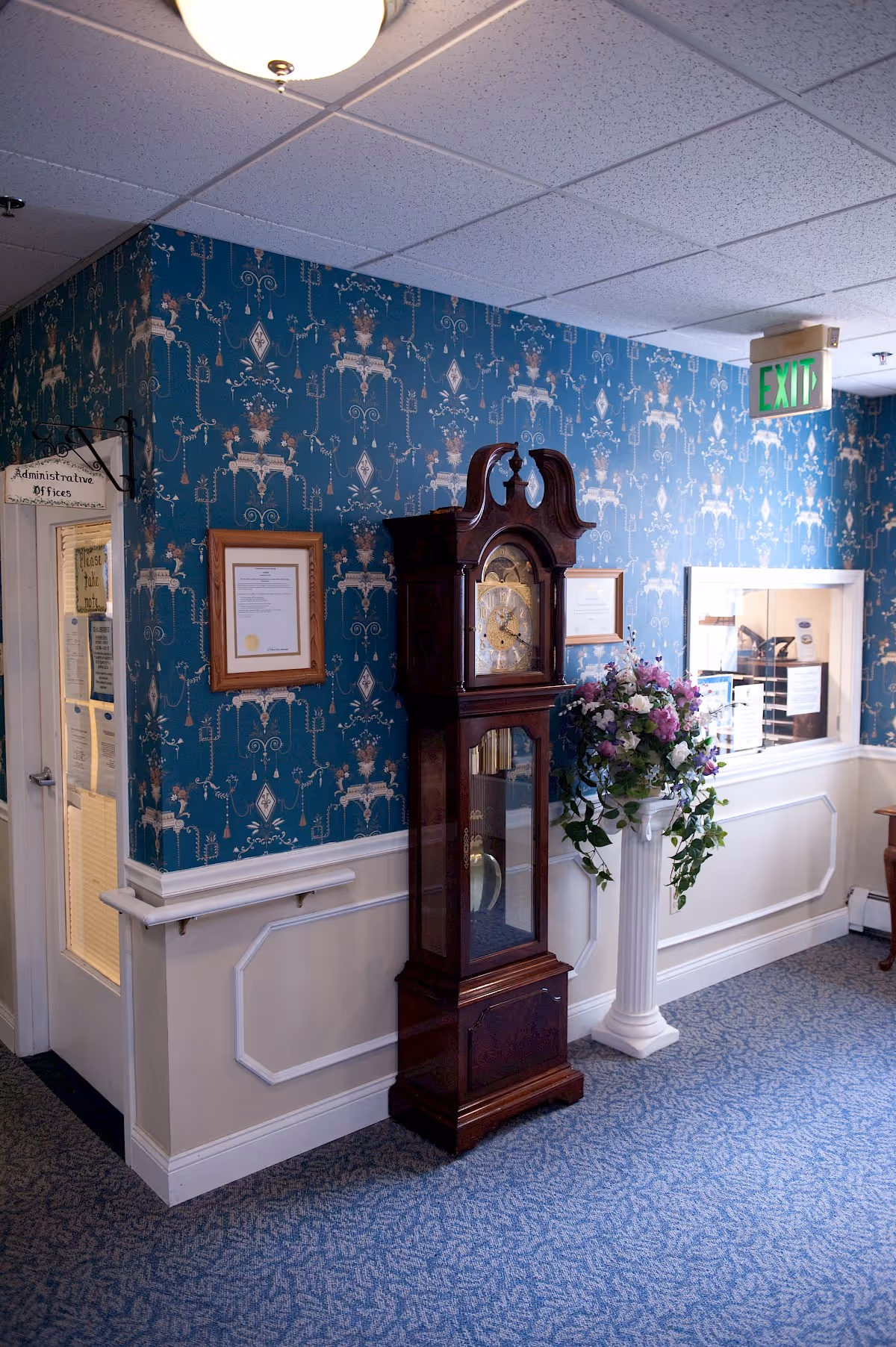 Interior hallway corner with blue patterned wallpaper and white wainscoting. A tall wooden grandfather clock stands next to a white pedestal holding a floral arrangement. There are framed documents on the wall and a door labeled 'Administrative Offices'. An exit sign is visible above a windowed reception area.