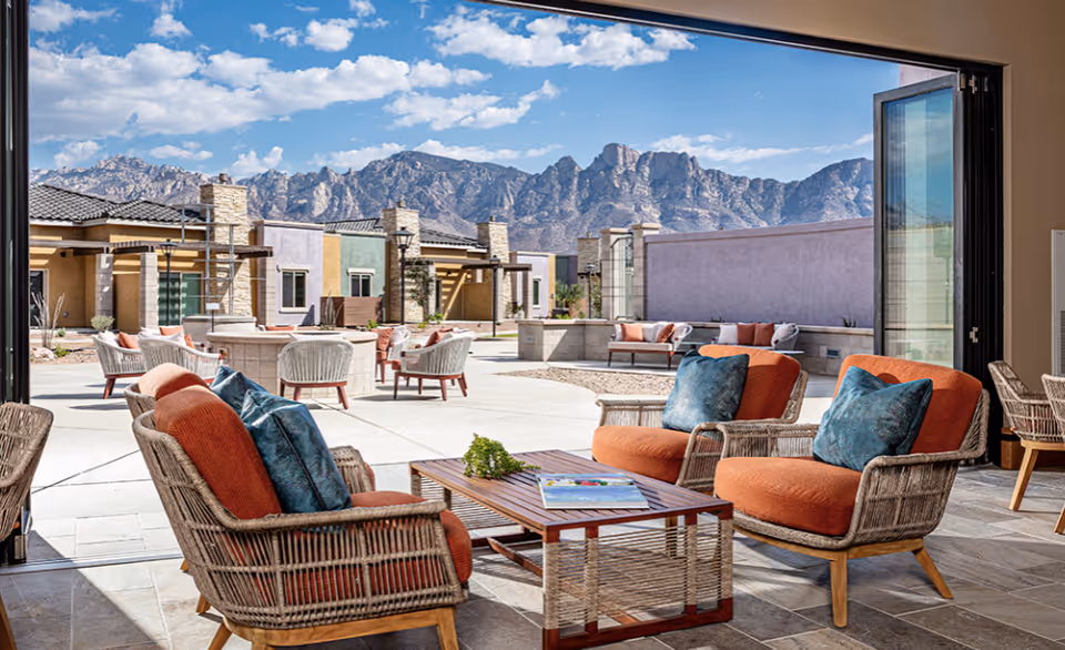 Wicker lounge chairs and a coffee table in an open indoor-outdoor seating area overlooking a sunny courtyard and mountain range.
