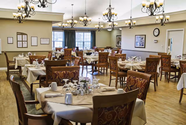 A spacious dining room with multiple round tables covered with beige tablecloths, each set with glassware, napkins, and silverware. The room features wooden chairs with patterned upholstery, large windows with dark curtains, and several ornate chandeliers hanging from the ceiling. The walls are decorated with framed pictures and a clock.