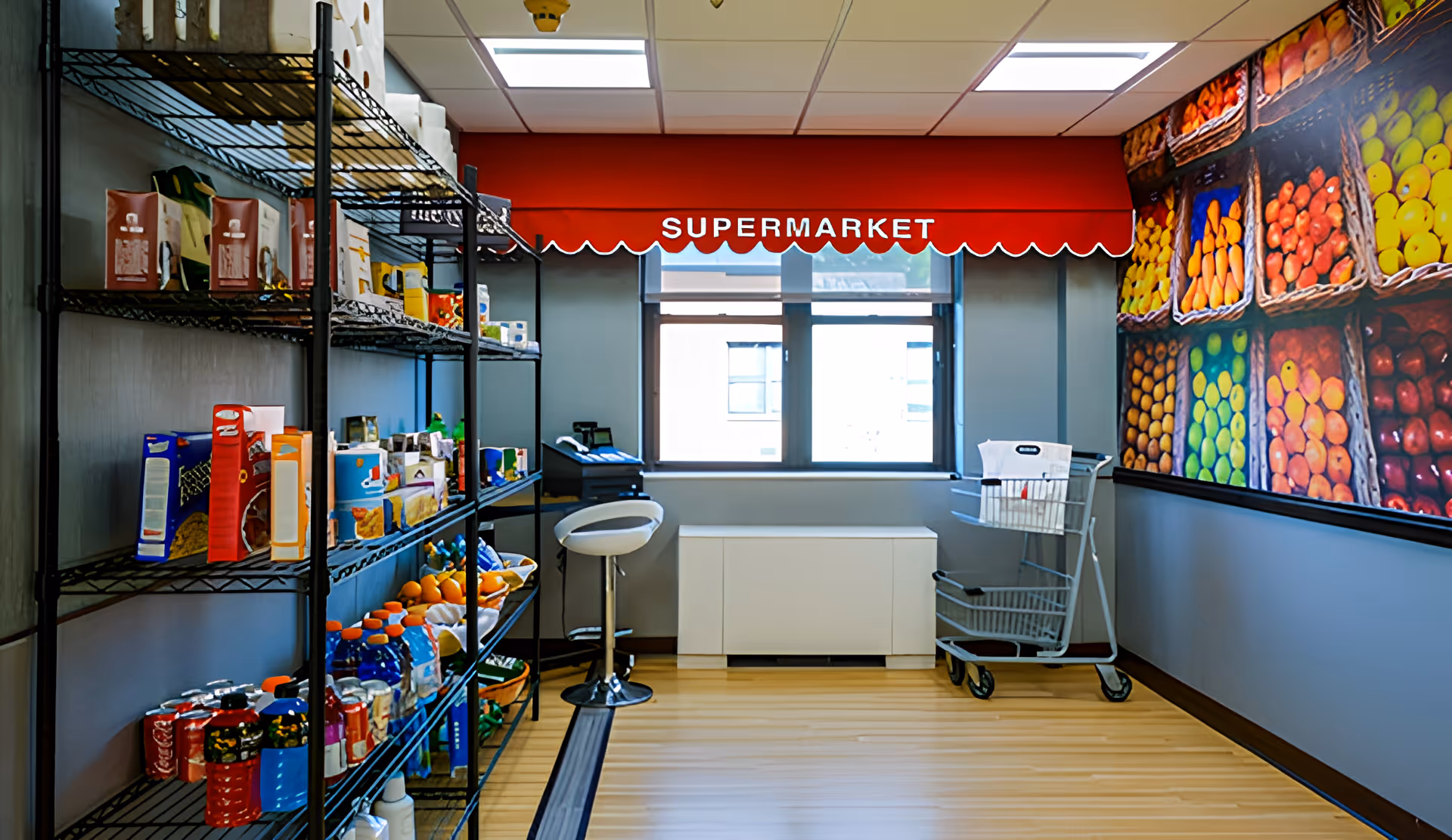 Interior of a small supermarket area with metal shelves stocked with various grocery items, a white stool, a shopping cart, and a large wall mural of colorful fruits and vegetables. A red awning with the word 'SUPERMARKET' is above a window letting in natural light.
