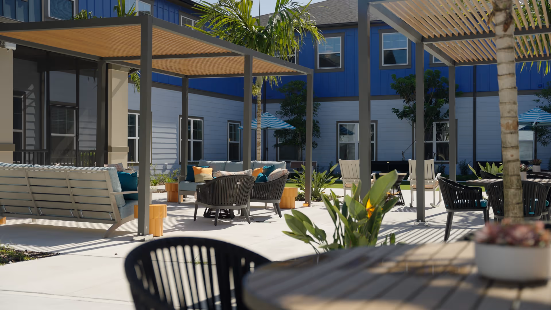 Outdoor seating area at Arya at DeBary with modern patio furniture including sofas, chairs, and tables under pergolas, surrounded by plants and a building with blue and white siding in the background.