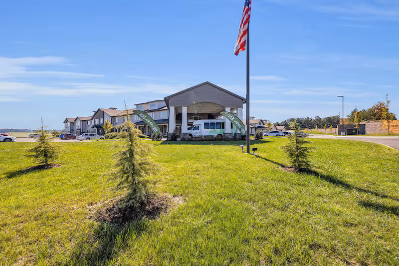 Front exterior view of Avail Senior Living building with a covered entrance, an American flag on a pole, several small trees planted in a grassy area, parked cars, and a clear blue sky.