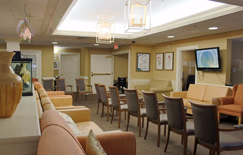 A spacious assisted living facility common area with multiple chairs arranged in rows facing a wall-mounted television. The room features beige walls, a variety of seating options including sofas and armchairs, framed artwork on the walls, and decorative ceiling lights. There is also a large vase and a fish tank on a counter to the left.