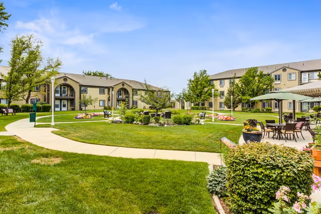 Outdoor view of Cedarwood at Sandy senior living facility showing a well-maintained green lawn with a curved concrete pathway, trees, flower beds, benches, and patio seating with tables and umbrellas. Two beige multi-story buildings with balconies and windows surround the garden area under a clear blue sky.