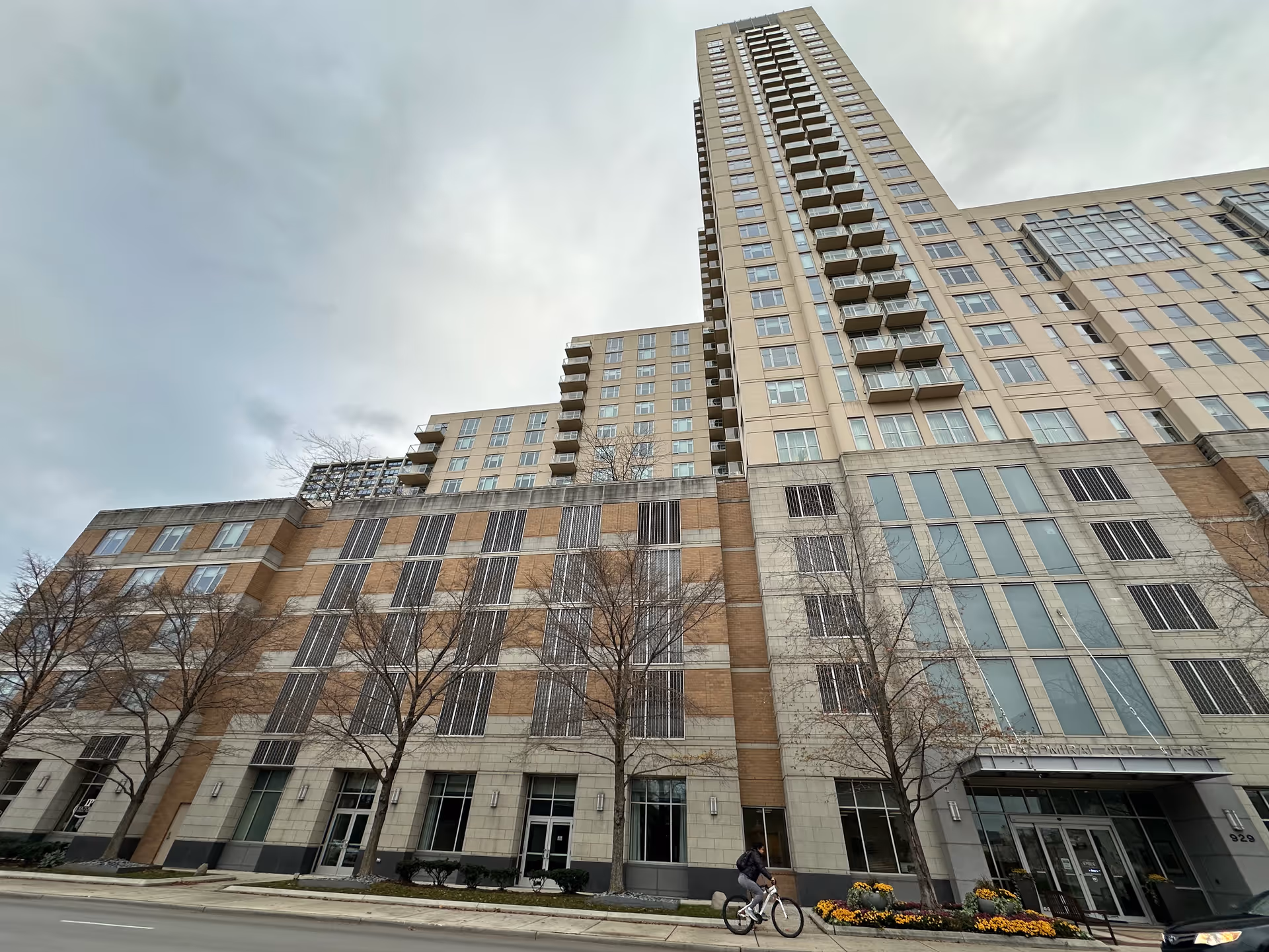 Exterior view of a tall multi-story residential building with balconies and large windows. The building has a mix of beige and light brown facade with some trees and flower beds in front. A person is riding a bicycle on the street in front of the building.