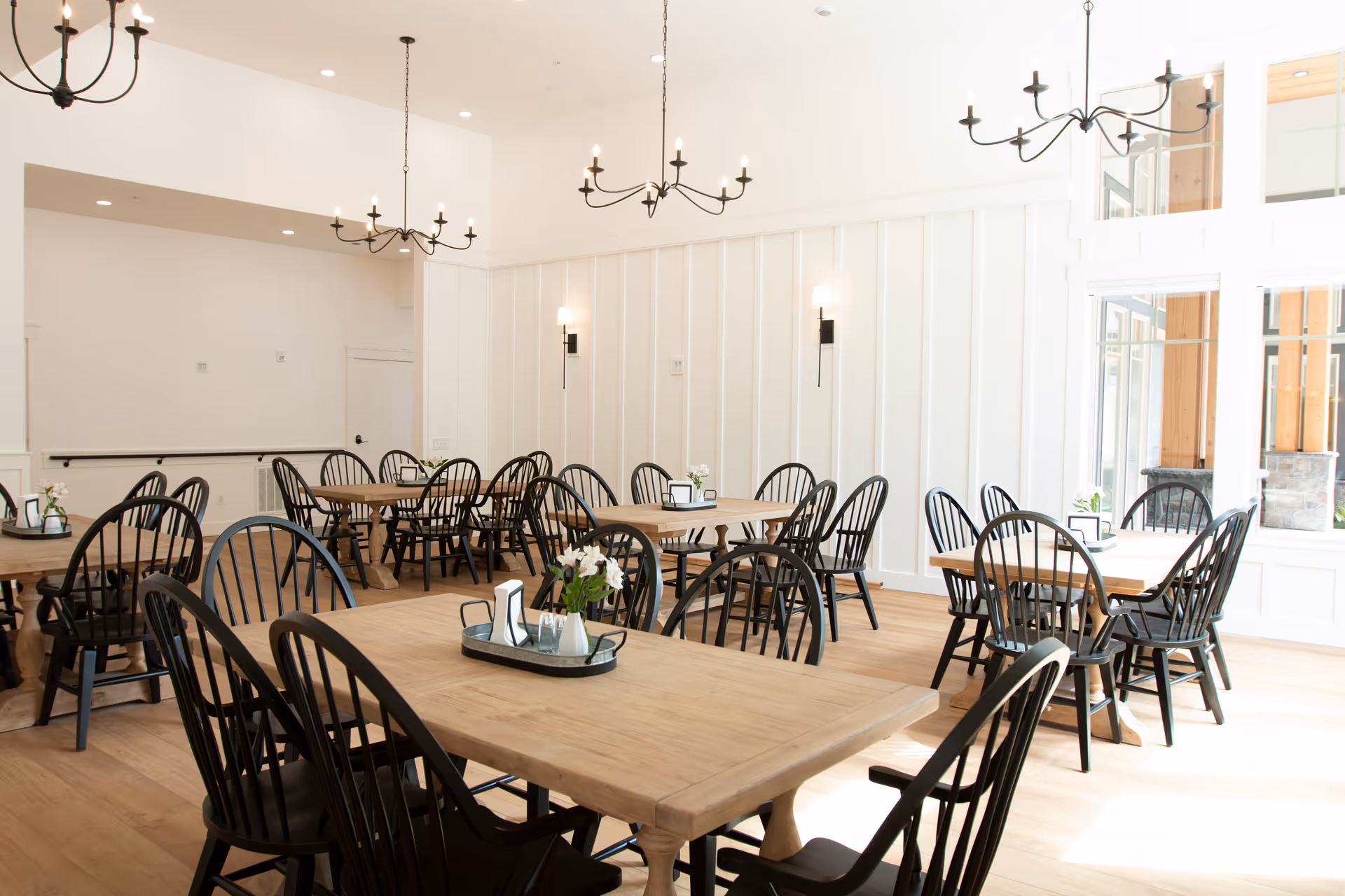 Bright dining room with multiple wooden tables and black chairs arranged neatly. The room features white paneled walls, large windows allowing natural light, and black chandeliers hanging from the ceiling. Each table has a small centerpiece with flowers and condiments.