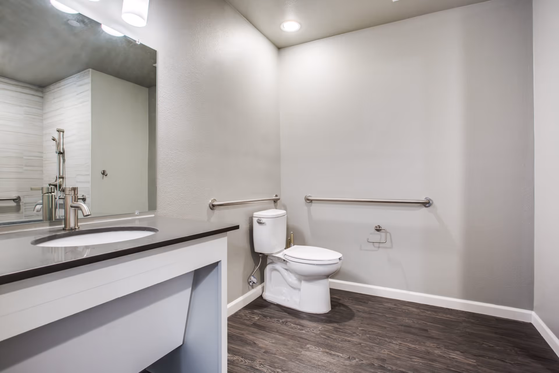 A modern bathroom with a white toilet, grab bars on the walls, a dark countertop with a sink, a large mirror, and wood-style flooring.
