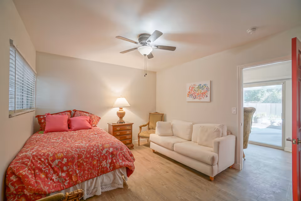 A cozy bedroom featuring a bed with red floral bedding and pink pillows, a wooden nightstand with a lamp, a beige armchair, and a white sofa. A ceiling fan with a light is mounted on the ceiling. There is a window with blinds on the left wall and an open door on the right leading to a bright outdoor patio area.