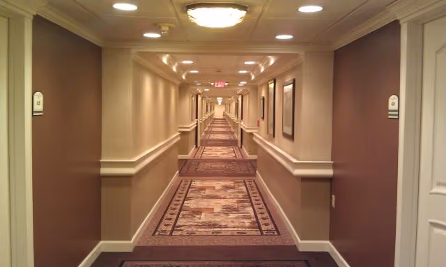 Long, well-lit hallway in a senior living facility with patterned carpet, beige walls, handrails on both sides, and multiple doors along the corridor. Ceiling lights and an exit sign are visible at the far end.