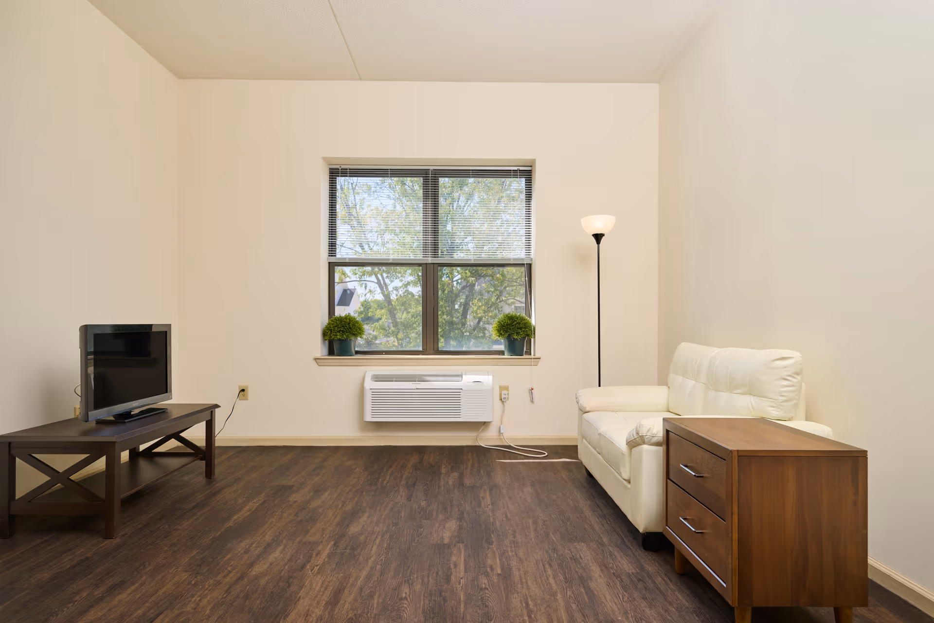 Small living room with a white sofa and wooden side table, TV on a low table, a window air conditioner, and a floor lamp.