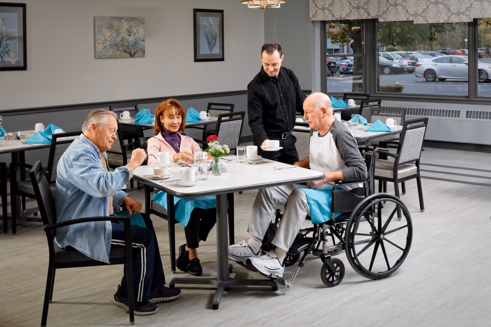 Three elderly individuals sitting around a dining table in a senior living facility dining room. One man is in a wheelchair, and a staff member is serving a cup of coffee to him. The table is set with cups, glasses, and a small flower arrangement. The room has large windows showing a parking lot outside, and tables with turquoise napkins are visible in the background.