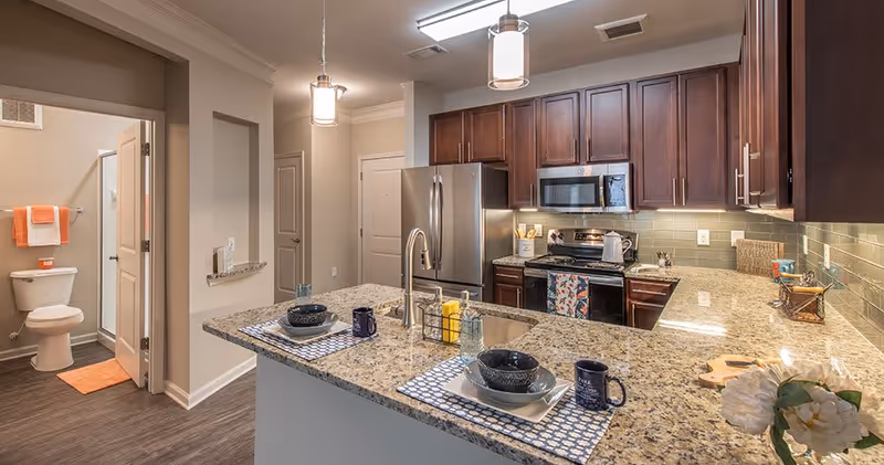 Modern kitchen with granite countertops, dark wood cabinets, stainless steel refrigerator, stove, and microwave. The kitchen island has two place settings with bowls, plates, mugs, and placemats. To the left, there is a view into a bathroom with a toilet, shower, and orange towels.