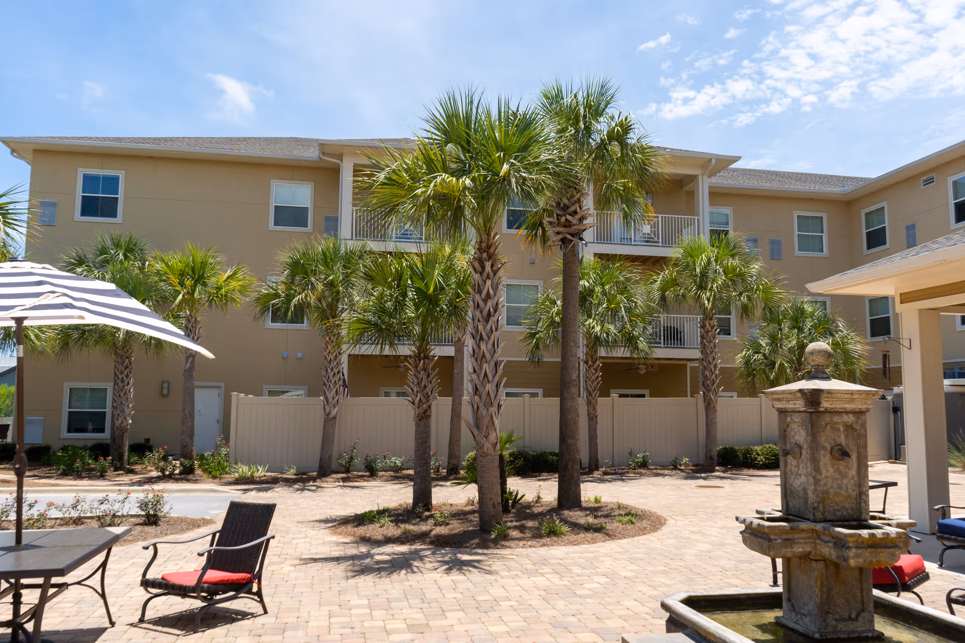 Outdoor courtyard area of a senior living facility with palm trees, a stone water fountain, patio chairs with red cushions, a table with a striped umbrella, and a three-story beige building in the background under a partly cloudy sky.