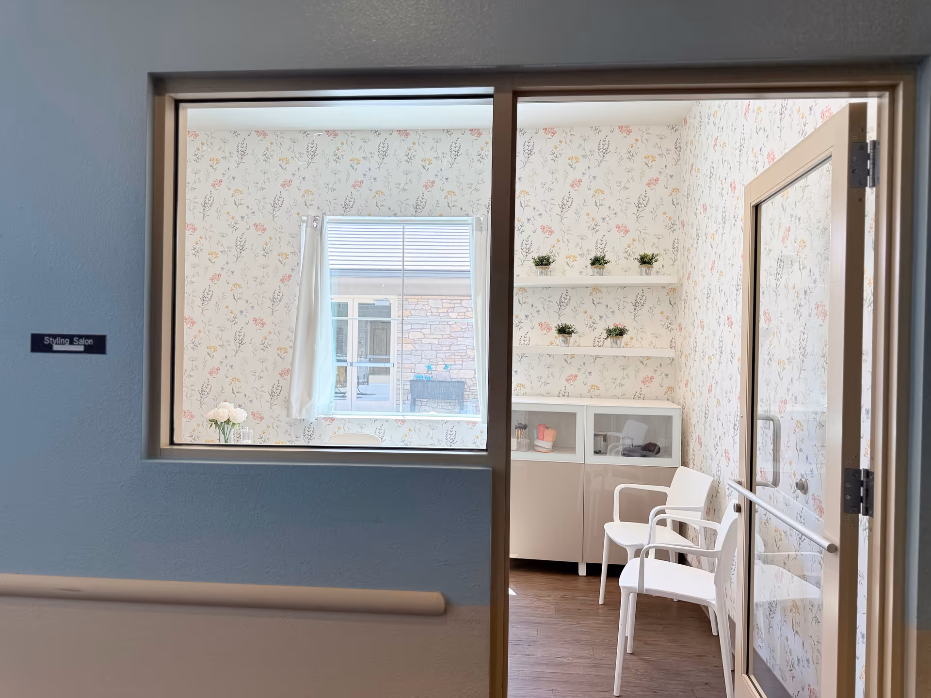 Interior view of a small styling salon room with floral wallpaper, two white chairs, a white cabinet with glass doors, and three shelves holding small potted plants. A window with white curtains looks out to an exterior wall with stone detailing.