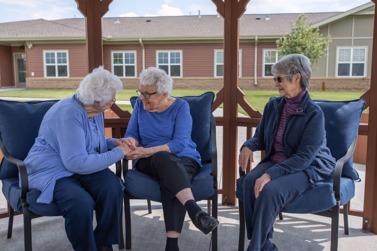 Three elderly women sitting on blue cushioned chairs under a wooden gazebo outside, engaged in conversation and holding hands, with a building and green lawn visible in the background.