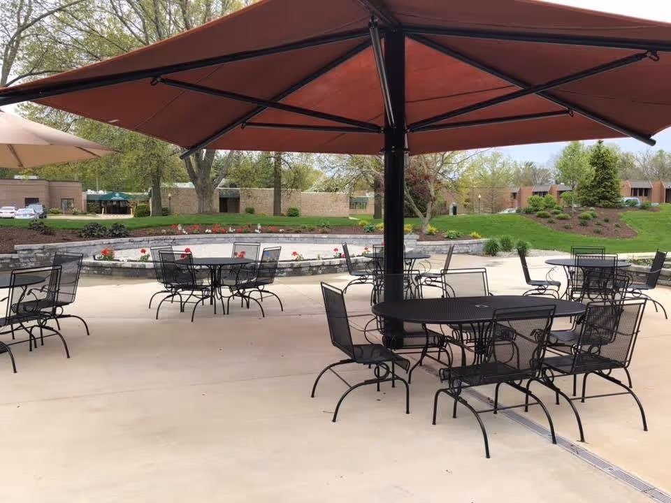 Outdoor patio area with multiple black metal tables and chairs under large umbrellas, surrounded by landscaped greenery and buildings in the background.
