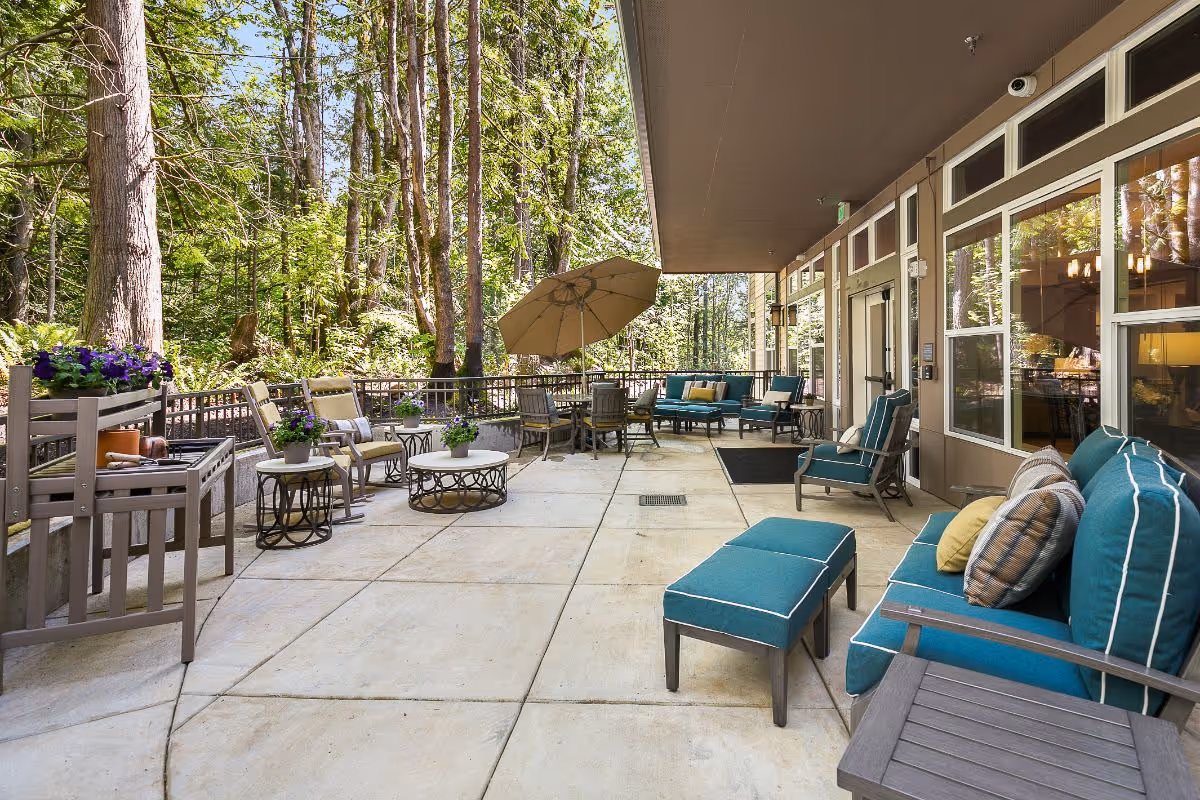 Outdoor patio area at Sunrise of Issaquah featuring cushioned seating, chairs, tables, potted plants, and a large umbrella, surrounded by trees and greenery.