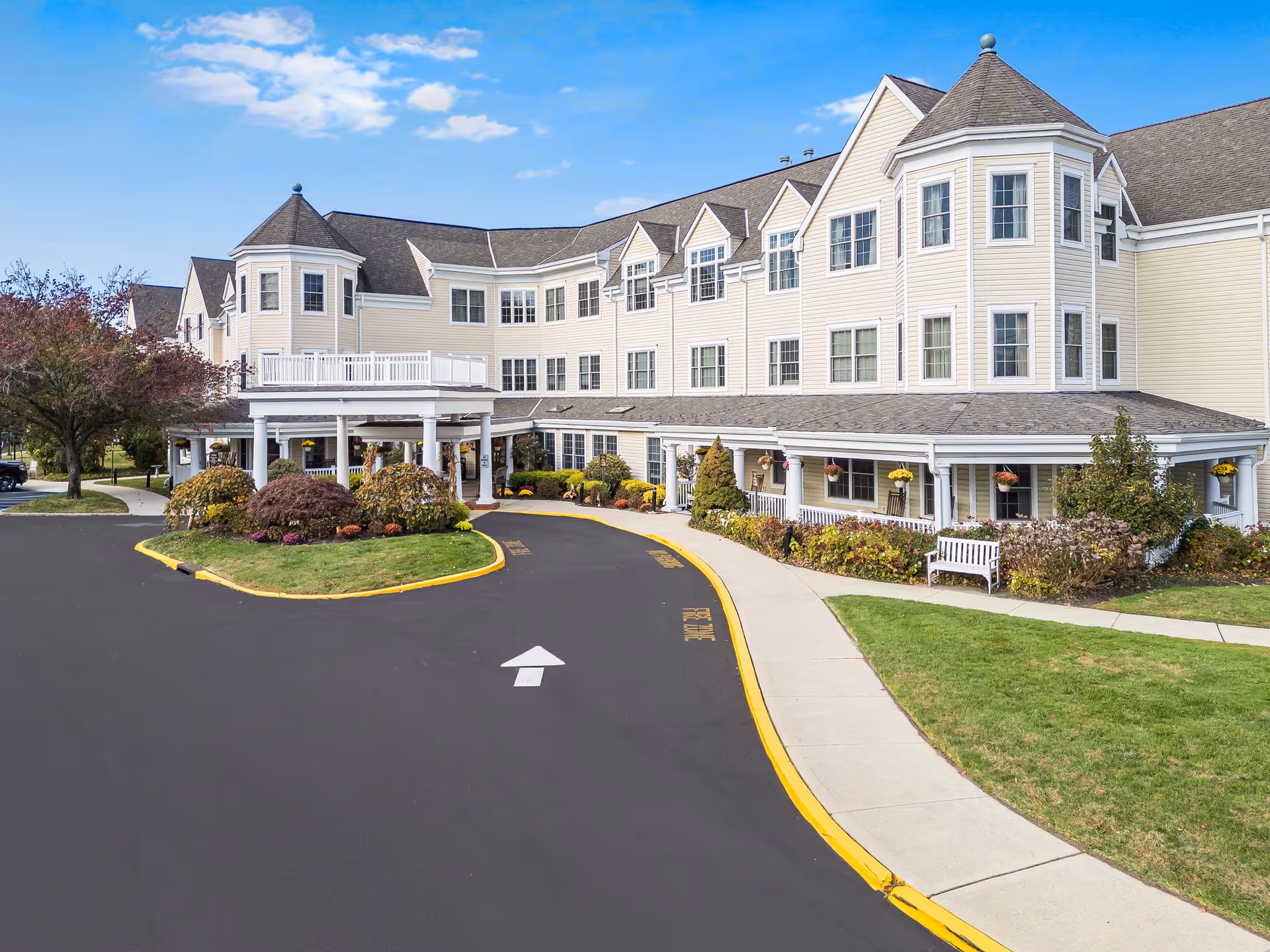 Exterior view of a large, light yellow senior living facility building with multiple windows and a covered entrance. The driveway curves around landscaped greenery with bushes and flowers. The sky is blue with a few clouds.