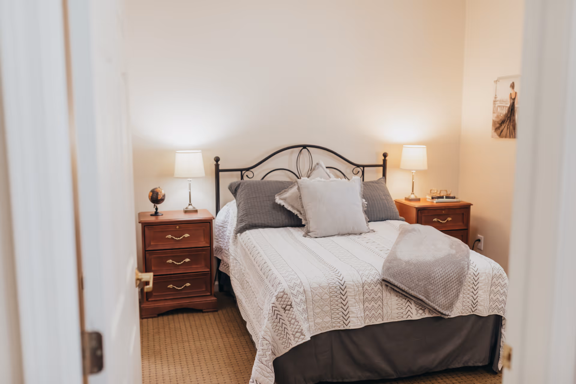 A cozy bedroom with a neatly made bed featuring a white and gray patterned quilt, multiple pillows, and a folded gray blanket at the foot. There are two wooden nightstands on either side of the bed, each with a lit table lamp. A small globe is on the left nightstand, and a framed picture hangs on the right wall. The room has beige walls and carpeted flooring.