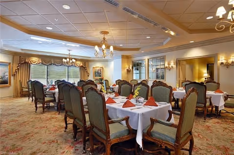 Elegant dining room with multiple round tables covered with white tablecloths and set with red folded napkins, fine china, and silverware. The room features ornate wooden chairs with green upholstery, chandeliers hanging from a white ceiling, large windows with draped curtains, and warm lighting from wall sconces.