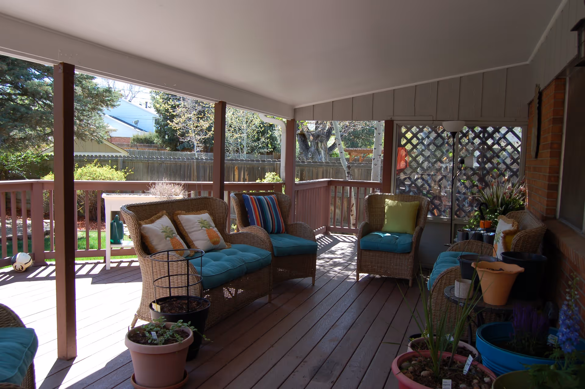 Covered outdoor patio area with wicker furniture including chairs and a loveseat with blue cushions and decorative pillows. Several potted plants are placed around the patio, and a wooden fence and trees are visible in the background.