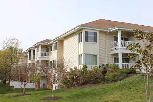 Exterior view of a multi-story residential building with beige siding, balconies, and a sloped roof, surrounded by green grass, small trees, and shrubs under a clear sky.