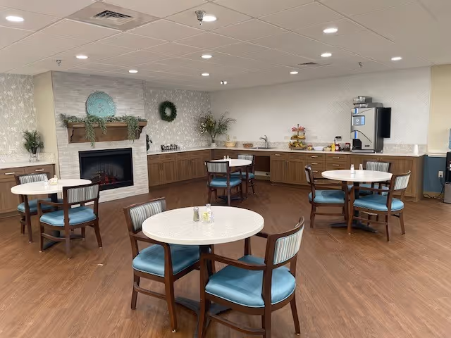 A cozy dining area with round tables and wooden chairs with blue cushions. The room features a fireplace with a decorative mantel, a wreath, and a large plate on the wall above it. There is a counter along the back wall with a coffee machine and various items, and the floor is wood with a light-colored ceiling and recessed lighting.