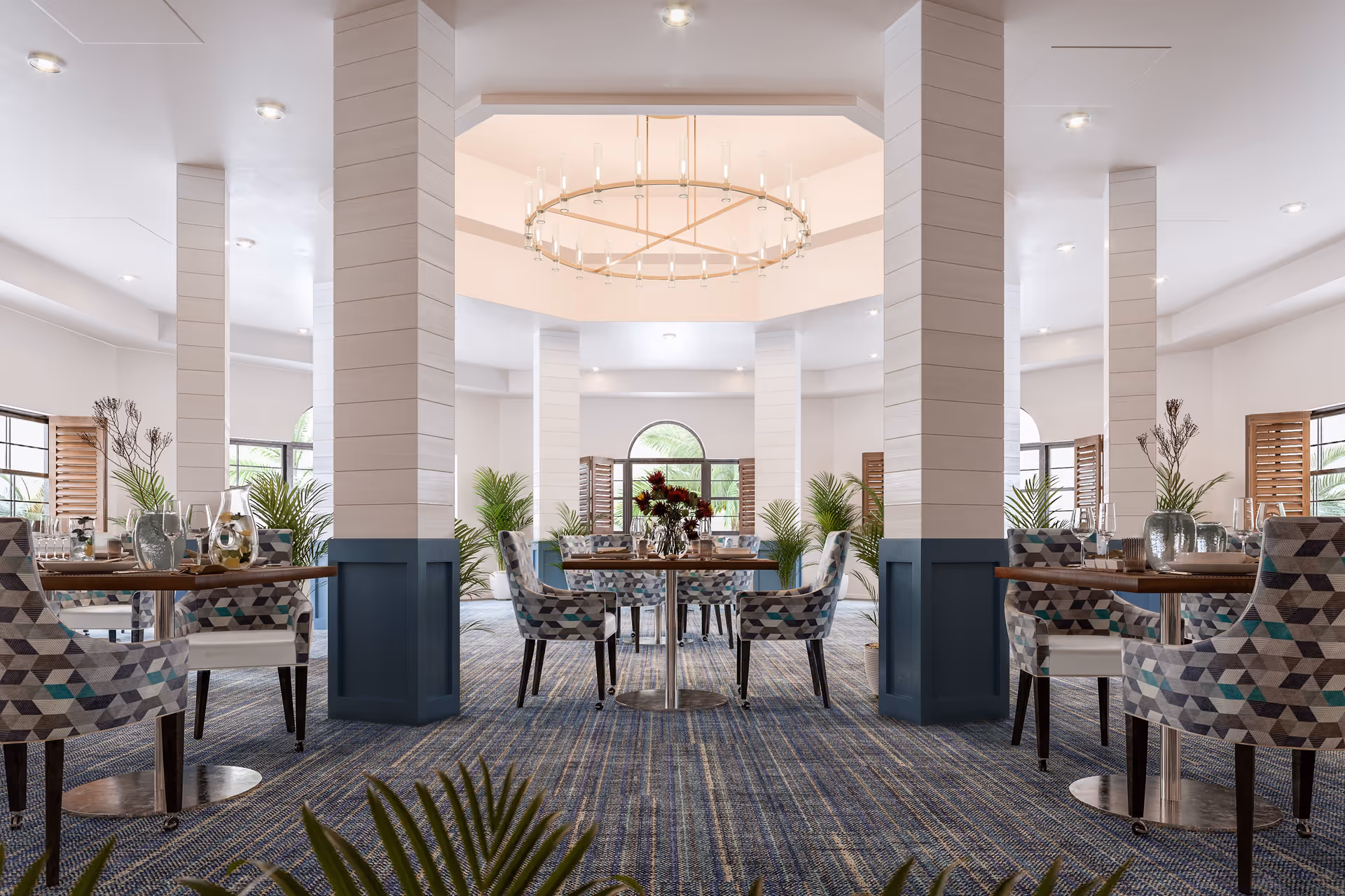 A bright and spacious dining room with patterned upholstered chairs around wooden tables. The room features multiple white pillars with blue paneling at the base, large windows with wooden shutters, green plants, and a large circular chandelier hanging from the ceiling.